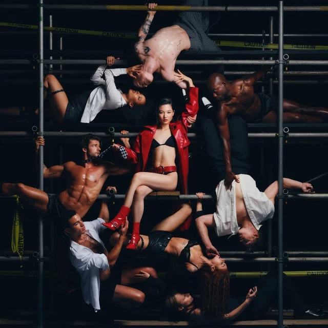 Chloe Qisha est au centre de la photo, en sous-vêtements avec une veste rouge en cuir et des bottines rouges en cuir. Elle est assise sur un échafaudage à plusieurs étages, entourée d'hommes et femmes plus ou moins dénudés, suspendus aux barres.